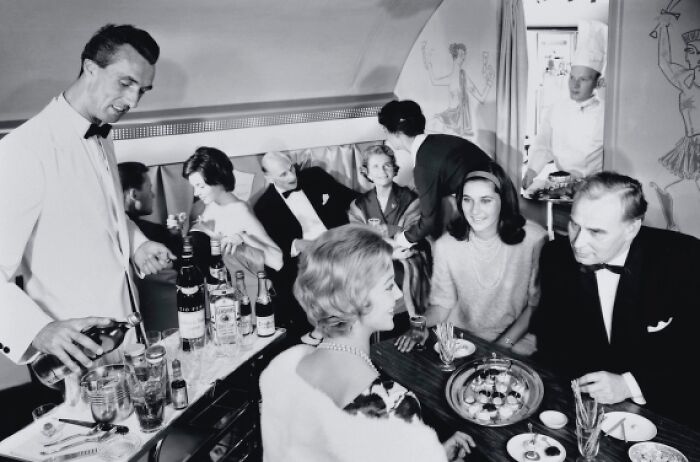Vintage black and white photo of glamorous passengers enjoying upscale in-flight service on a luxury airplane.