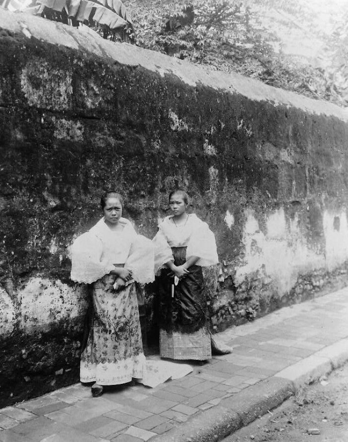 Two Filipino women in traditional attire standing by an old stone wall in the Philippines in the 1890s.