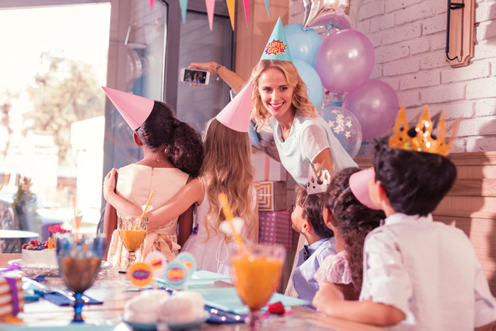 Woman at niece's birthday party taking selfie with children wearing party hats celebrating together indoors. Woman at niece's birthday party taking selfie with children wearing party hats celebrating together indoors.