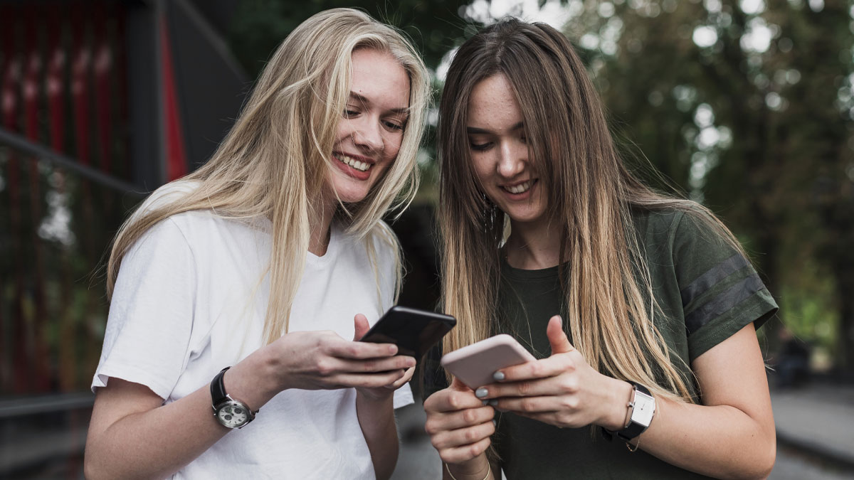 Two young women smiling and looking at their phones, enjoying funny and cute animal memes together outdoors.