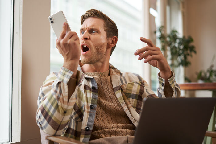 Man upset and shocked looking at phone while sitting by laptop indoors, reacting to photos of his ex at sister’s BBQ. Man upset and shocked looking at phone while sitting by laptop indoors, reacting to photos of his ex at sister’s BBQ.