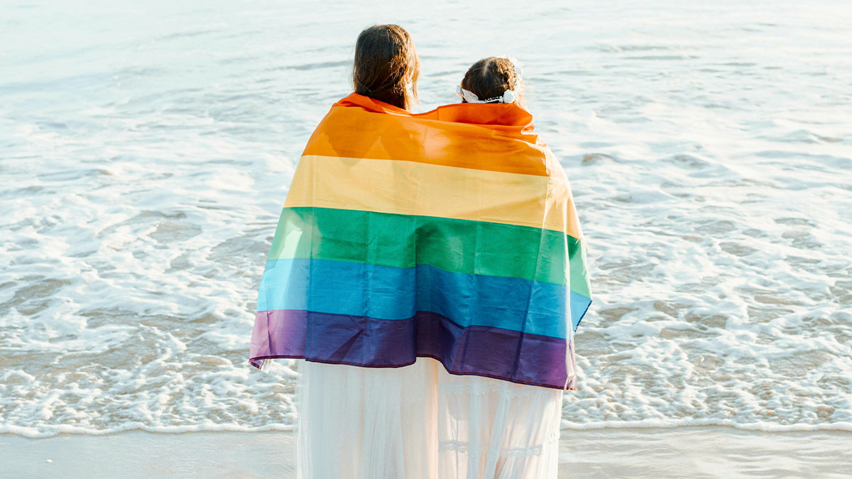 Woman and child wrapped in rainbow flag standing by the ocean, symbolizing womanu2019s fight with stepsister and family ties.