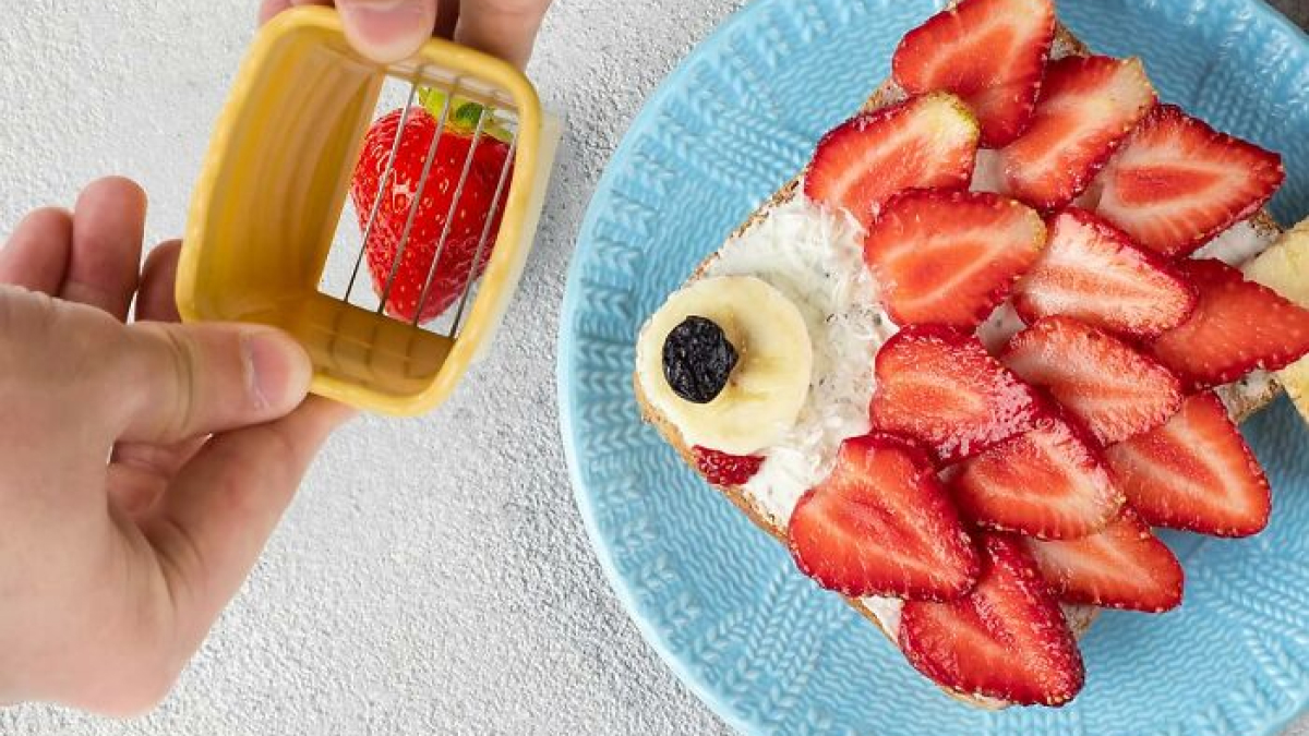 Hand holding a kitchen problem-solver tool slicing strawberries for a fruit-topped toast on a blue plate.