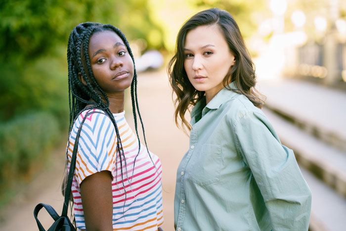 Two teenage girls, one black and one Asian, standing outdoors with serious expressions, highlighting a tense moment. Two teenage girls, one black and one Asian, standing outdoors with serious expressions, highlighting a tense moment.