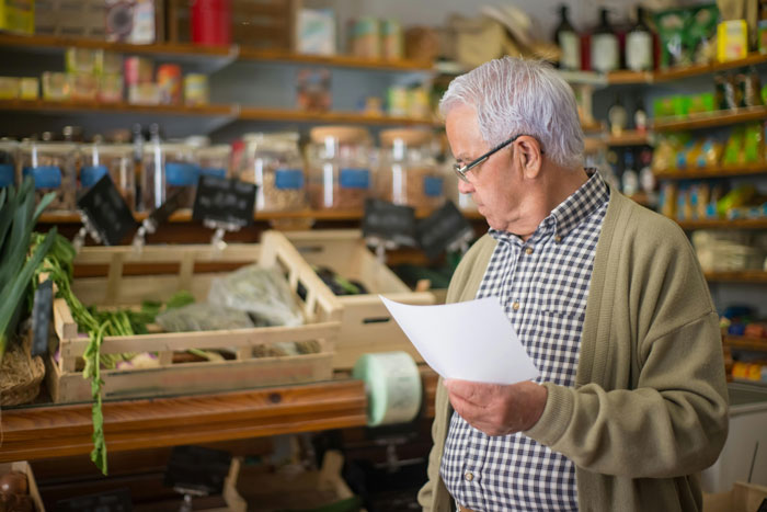Elderly man shopping at grocery store holding list, standing near fresh produce and shelves with packaged goods. Elderly man shopping at grocery store holding list, standing near fresh produce and shelves with packaged goods.