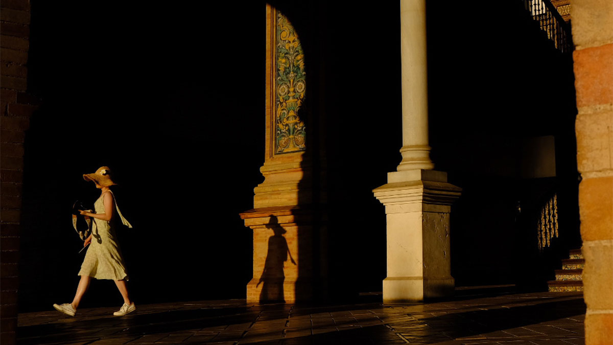 Woman in a sunhat walking on a street with strong shadows and architectural columns in a street photography moment