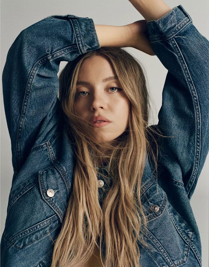 Young woman in denim jacket with arms raised, styled hair, and neutral expression illustrating Lizzo Sydney Sweeney parody topic. Young woman in denim jacket with arms raised, styled hair, and neutral expression illustrating Lizzo Sydney Sweeney parody topic.