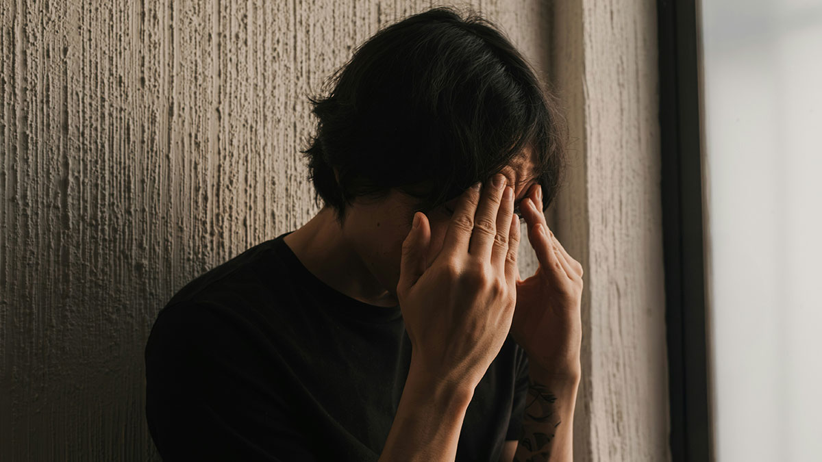 Teen with dark hair covering face with hands sitting by textured wall showing distress over father last name choice.