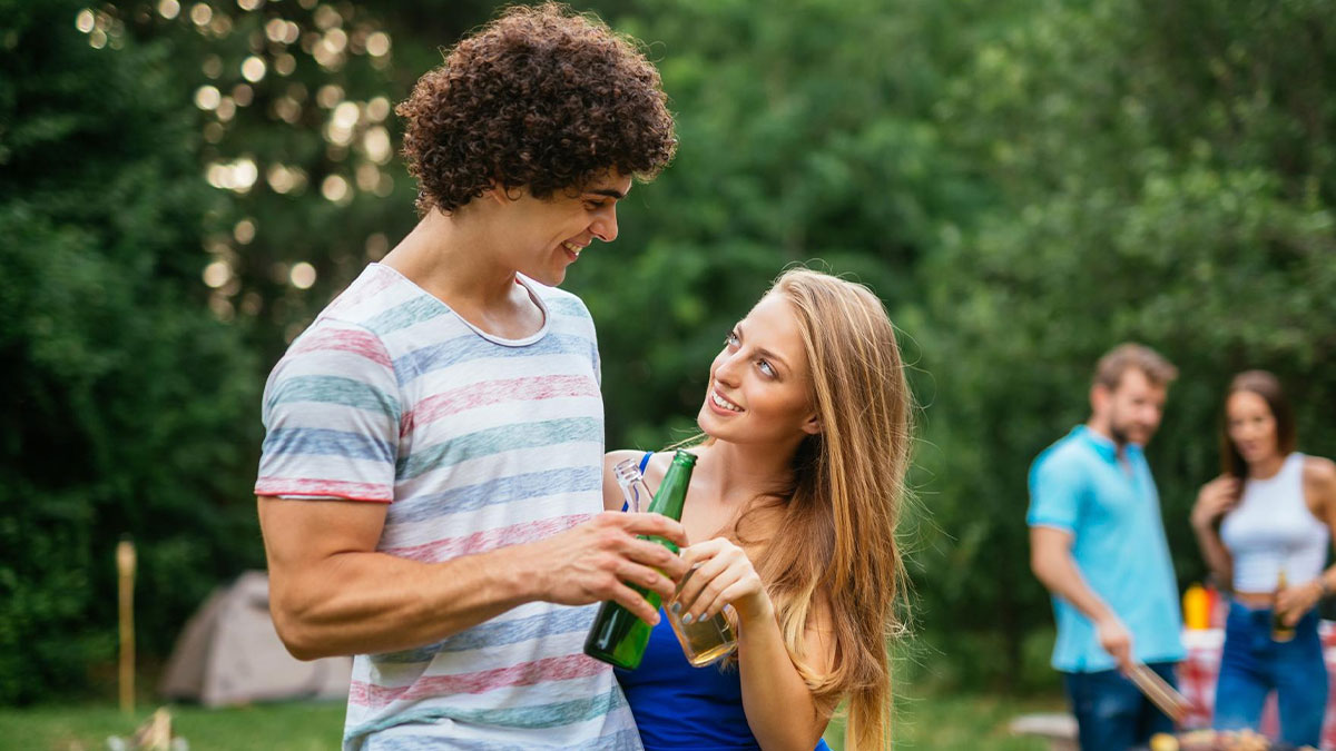 Young couple holding drinks and smiling during an outdoor gathering, with a barbecue in the background.