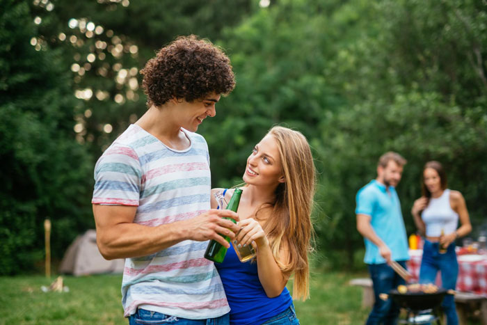 Young couple sharing drinks outdoors, while others barbecue in the background, reflecting on sister's boyfriend seeming quite fake. Young couple sharing drinks outdoors, while others barbecue in the background, reflecting on sister's boyfriend seeming quite fake.