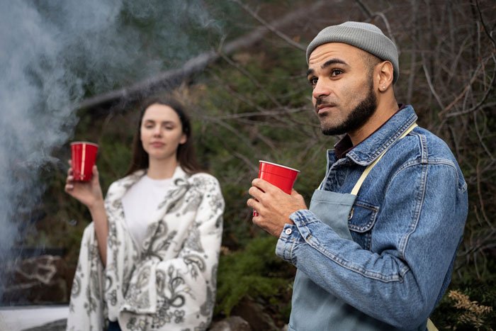 Person in denim jacket and beanie holding a red cup, thinking sister's boyfriend is quite fake outdoors near a fire. Person in denim jacket and beanie holding a red cup, thinking sister's boyfriend is quite fake outdoors near a fire.