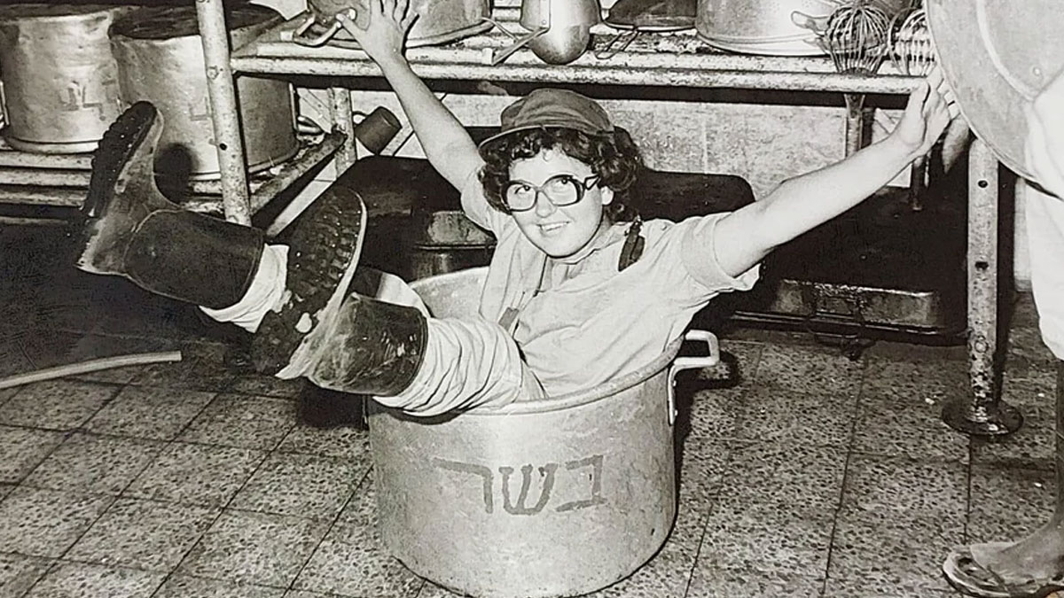 Young woman in boots and glasses playfully sitting inside a large cooking pot in a candid glimpse into the past.