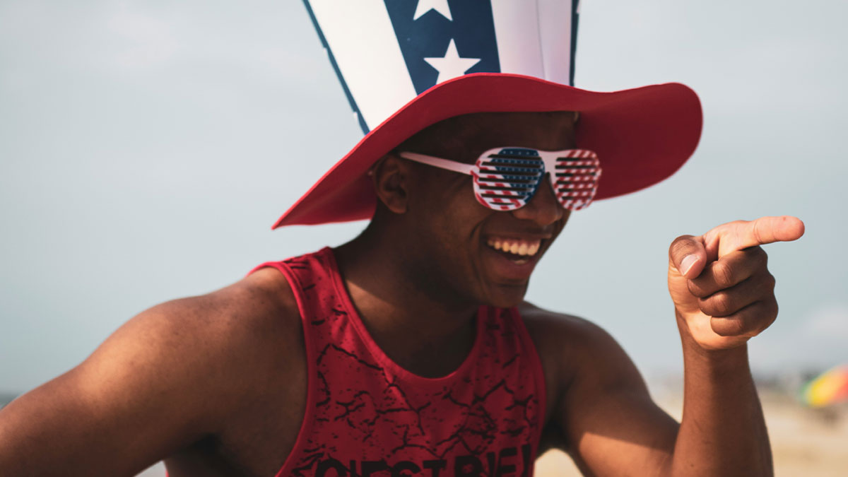 Man wearing patriotic American flag hat and sunglasses smiling and pointing, showing things Americans do abroad.