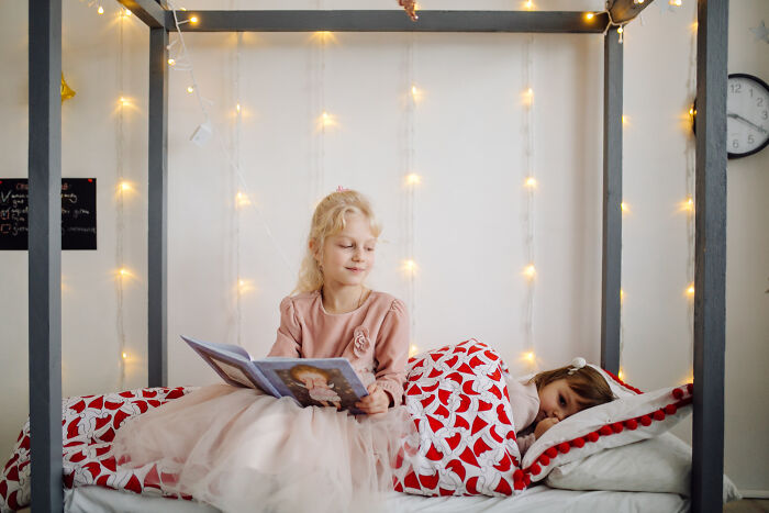 Two children in a cozy bedroom with fairy lights, one reading a book while the other sleeps, evoking childhood memories.