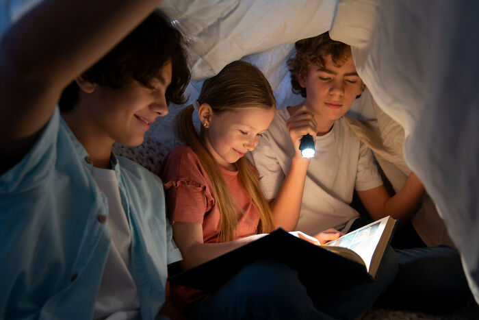 Three kids reading a book under a blanket with a flashlight, evoking childhood memories that felt sweet back then.