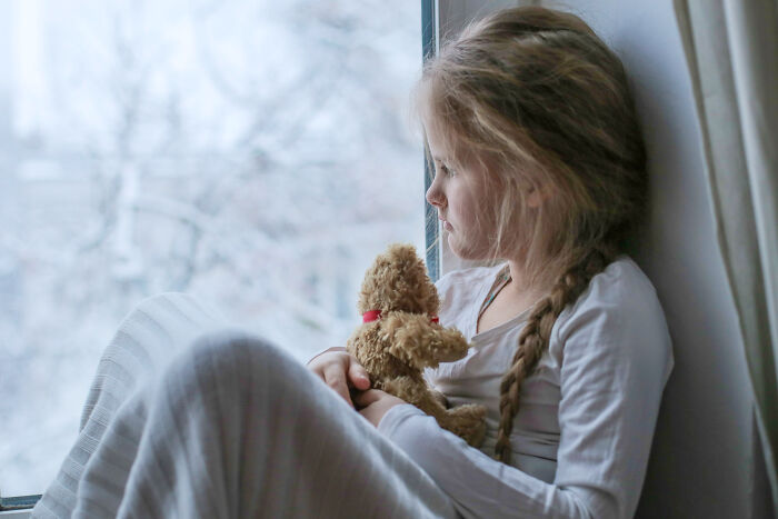 Young girl holding a teddy bear and looking out a window, evoking childhood memories that feel sweet but unsettling in retrospect.