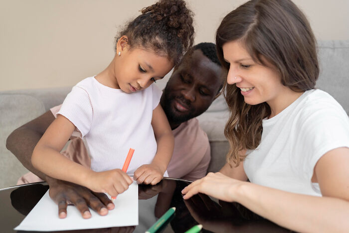 A young girl drawing with colored pencils while her parents watch closely, evoking childhood memories and family moments.
