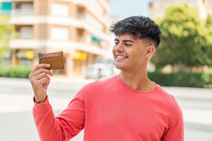 Young man smiling and holding a wallet outdoors, illustrating common tourist traps related to money and spending. Young man smiling and holding a wallet outdoors, illustrating common tourist traps related to money and spending.