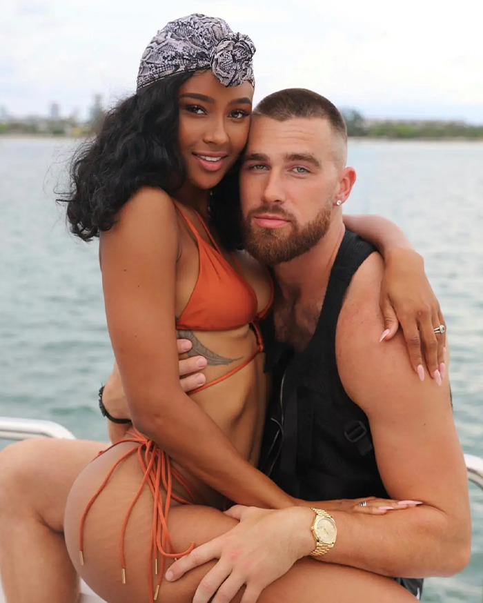 Travis Kelce's ex wearing a headscarf and bikini, embracing a man with a beard while sitting by the water. Travis Kelce's ex wearing a headscarf and bikini, embracing a man with a beard while sitting by the water.