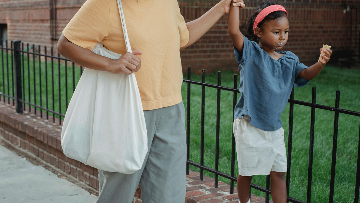 Adult holding hand of child eating while walking near black fence and brick wall outdoors in casual clothing