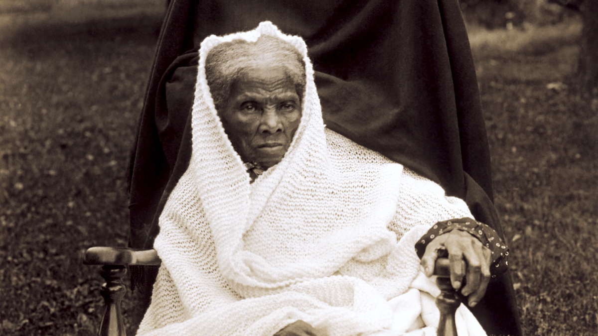 Elderly woman wrapped in a blanket sitting outdoors in a historic setting related to Americau2019s architectural heritage.