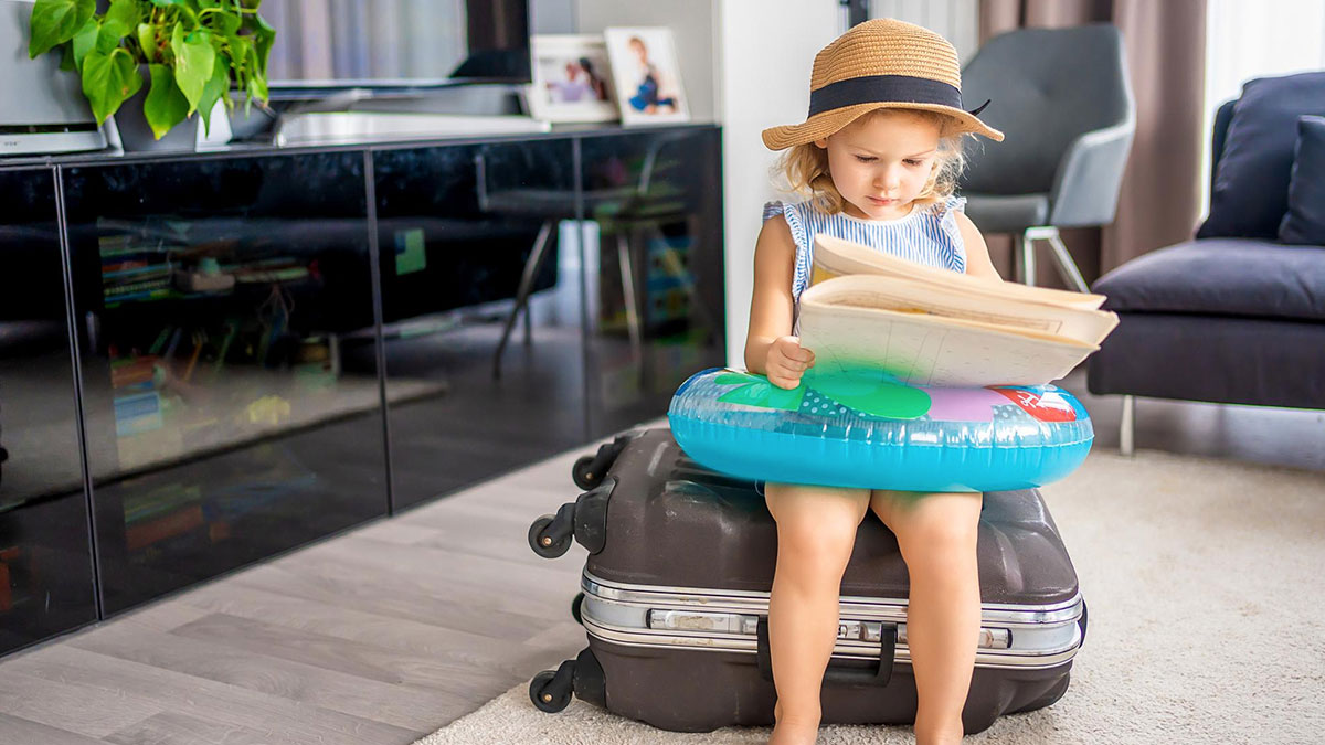 Toddler with inflatable ring and hat sitting on suitcase reading book in living room suggesting kid-free vacation conflict.
