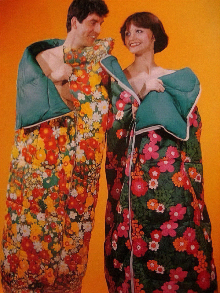 Couple wearing brightly colored vintage floral sleeping bags against an orange background, evoking old-timey style.