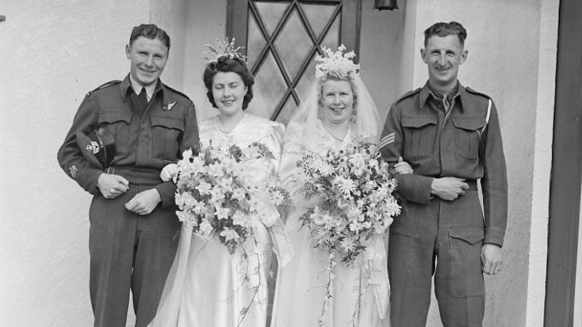 Two 1940s brides in wedding dresses holding large bouquets standing with grooms in military uniforms after wartime wedding.