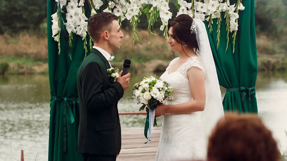 Groom holding microphone and bride with bouquet standing under floral arch at outdoor wedding ceremony