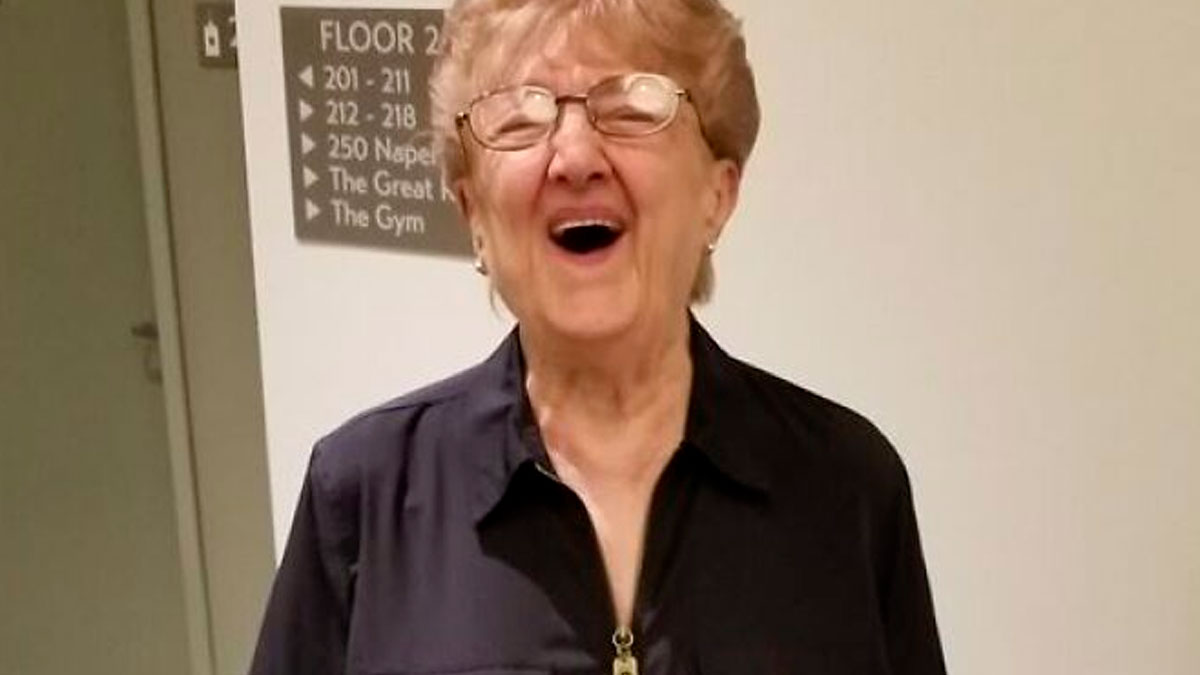 Elderly woman with glasses smiling warmly in a hallway, embodying the spirit of wholesome kind neighbors.