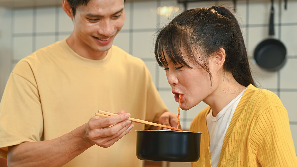 Couple in kitchen with woman tasting food from a pot held by husband, reflecting jealousy and memory of friendu2019s favorite drink.