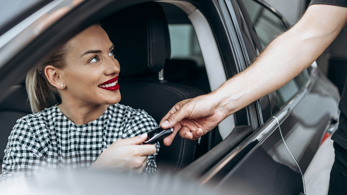 Wife sitting in car smiling as husband reaches in to take car key from her at the dealership.