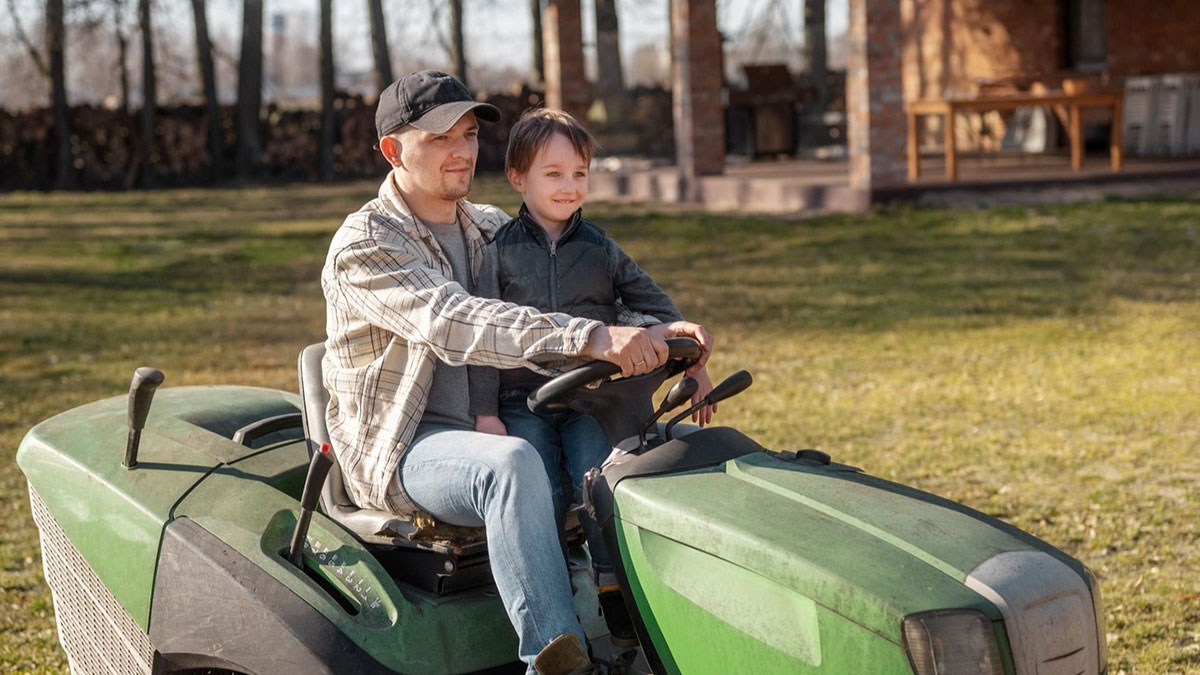 Man and school kid confidently riding a green lawn tractor on a sunny day in a backyard setting.