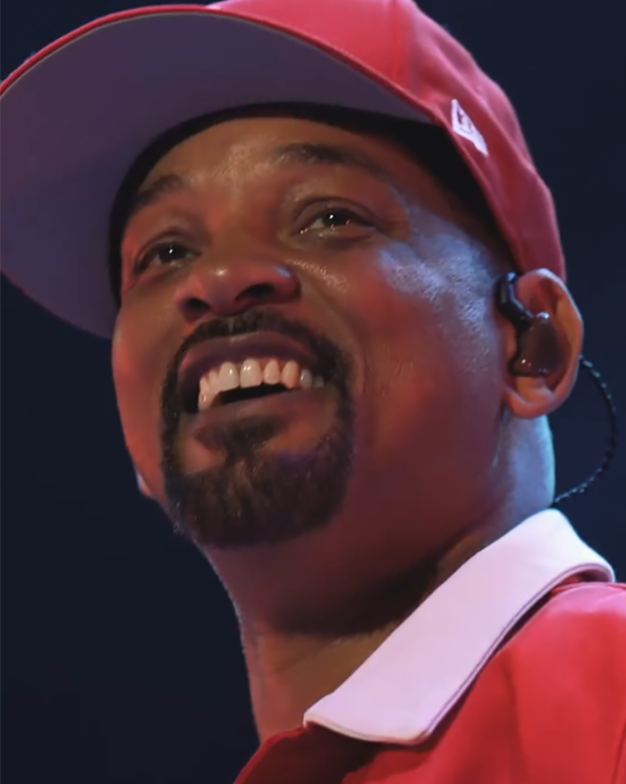 Will Smith smiling on stage wearing a red cap and shirt, related to being mocked for faking crowds with AI. Will Smith smiling on stage wearing a red cap and shirt, related to being mocked for faking crowds with AI.