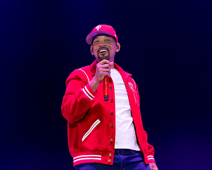Will Smith on stage wearing a red jacket and cap, holding a microphone, amid AI crowd manipulation mockery. Will Smith on stage wearing a red jacket and cap, holding a microphone, amid AI crowd manipulation mockery.