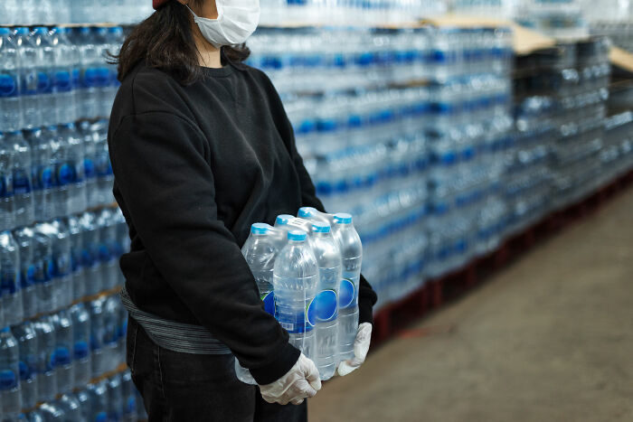 Person wearing a mask and gloves holding bottled water in a warehouse, illustrating unexpected things witnessed in strangers' homes.