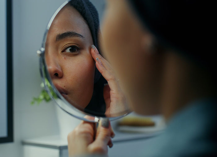 Woman examining her face in a handheld mirror, reflecting concerns related to the Love Island face trend and filler blindness. Woman examining her face in a handheld mirror, reflecting concerns related to the Love Island face trend and filler blindness.