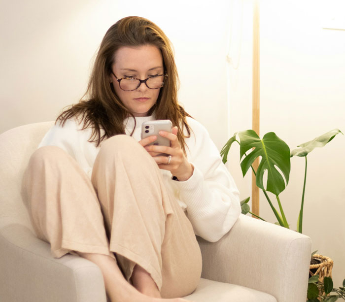 Woman sitting in chair focused on phone, reflecting on weight loss and reconnecting with good friend after years apart Woman sitting in chair focused on phone, reflecting on weight loss and reconnecting with good friend after years apart