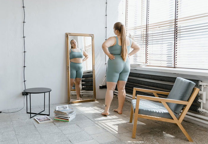 Woman in athletic wear standing in front of mirror, reflecting on weight and body image in a sunlit room. Woman in athletic wear standing in front of mirror, reflecting on weight and body image in a sunlit room.