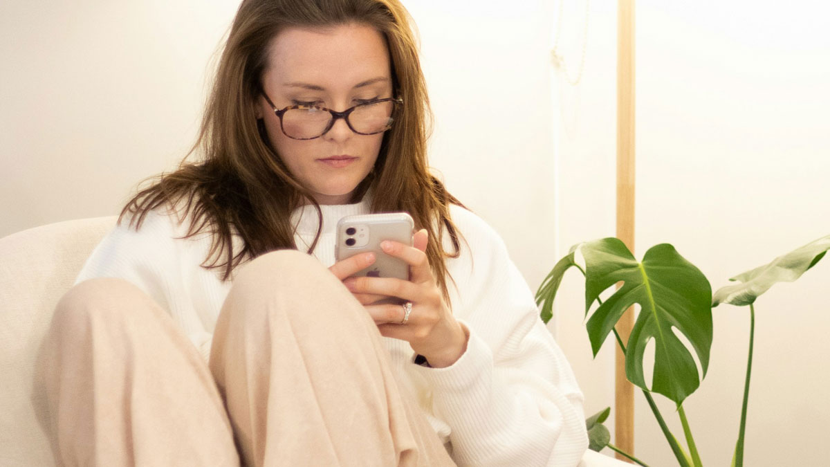 Woman with glasses sitting indoors looking at phone, reflecting on a good friend and weight loss conversation.