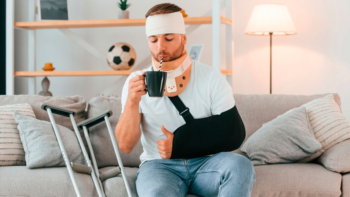 Man with injury wearing neck brace and arm sling, sitting on couch with crutches nearby, drinking from a mug.