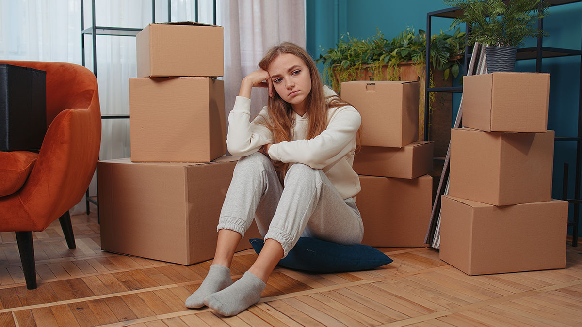Sad young woman sitting on floor surrounded by moving boxes, feeling needy and heartbroken after moving out from parents' home.