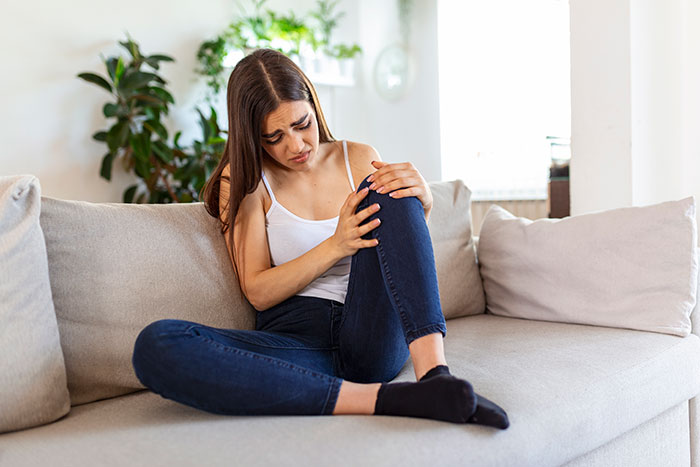 Young woman sitting on a couch holding her knee in pain, illustrating chronic pain dismissed by doctors until validated by husband. Young woman sitting on a couch holding her knee in pain, illustrating chronic pain dismissed by doctors until validated by husband.