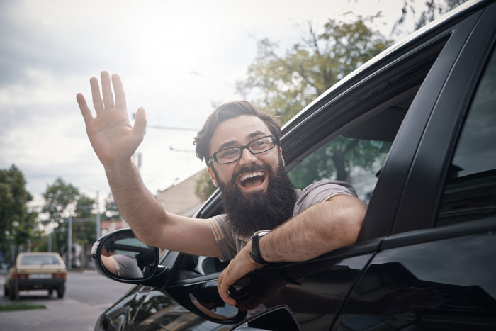 Man with glasses and beard happily waving from the driver’s seat of a black car, outside on a bright day.