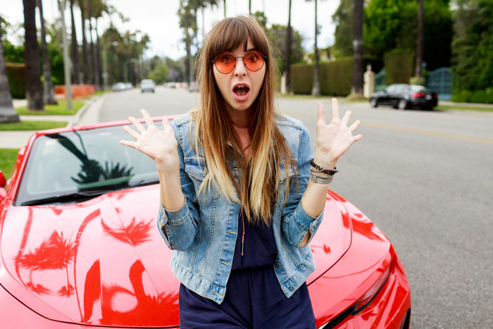 Woman in sunglasses and denim jacket reacting with surprise in front of a red car, illustrating lady in brand new Lexus scenario.