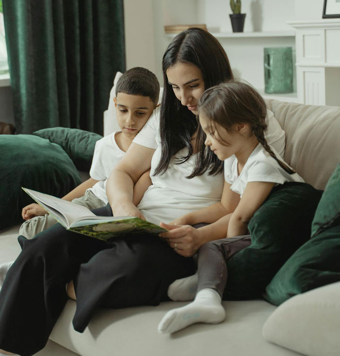 Woman reading a book with two children on a couch, highlighting challenges of babysitting her autistic nephew. Woman reading a book with two children on a couch, highlighting challenges of babysitting her autistic nephew.