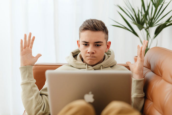 Teen boy sitting on couch with raised hands, looking frustrated at a laptop, related to autistic nephew babysitting issue. Teen boy sitting on couch with raised hands, looking frustrated at a laptop, related to autistic nephew babysitting issue.
