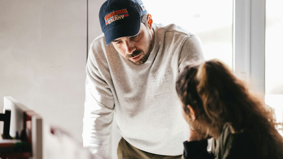 Man wearing a cap and gray sweatshirt talking seriously to a woman in a bright room, suggesting a hidden affair discussion.