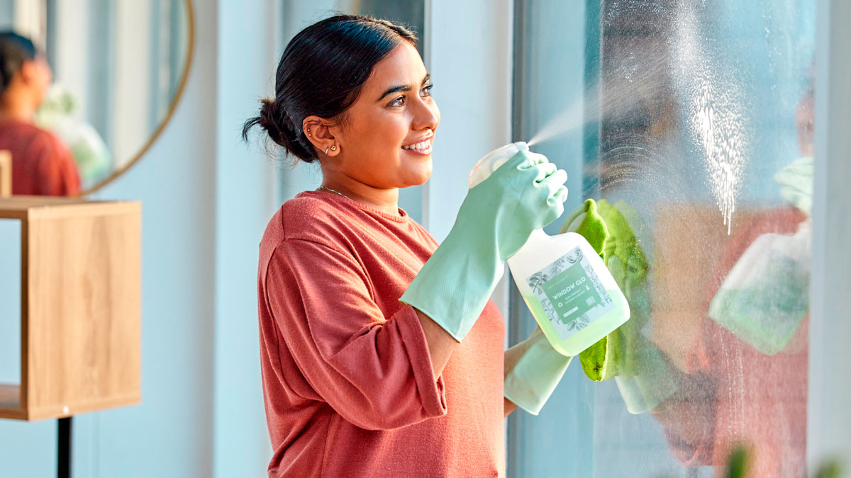 Woman wearing gloves cleaning a window with spray bottle, illustrating the challenge of maintaining a clean toddler environment.