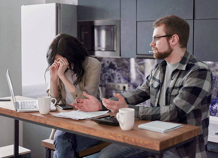 Woman feels financially trapped as jobless fiancé argues at kitchen table with bills and laptop nearby. Woman feels financially trapped as jobless fiancé argues at kitchen table with bills and laptop nearby.